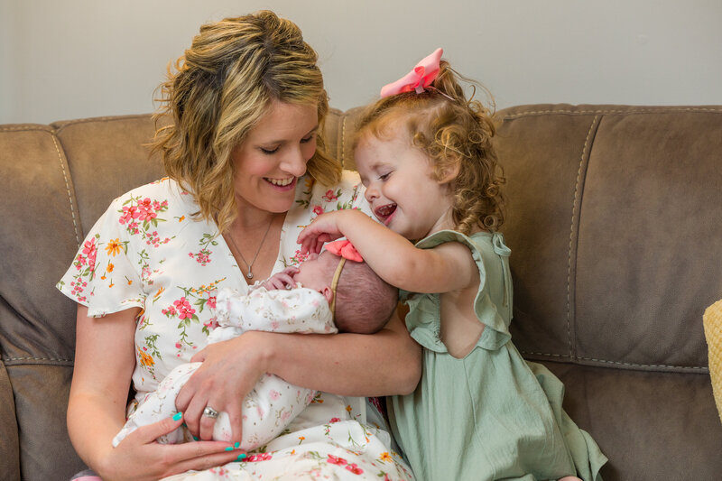 A little girl playfully points to her newborn sibling’s nose as their mom laughs in a heartwarming moment during a family session near Phoenixville.