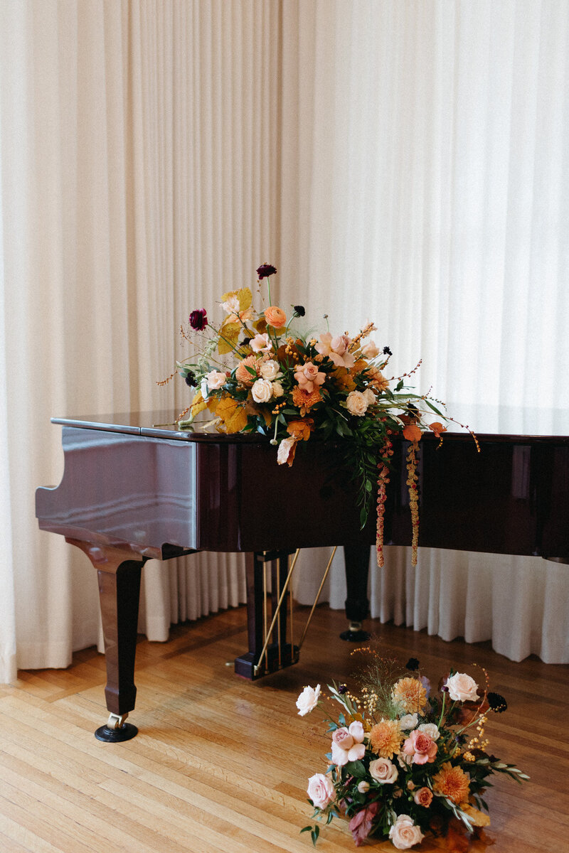Grand piano adorned with a floral arrangement, featuring roses and greenery, set against white curtains—a perfect scene for a Capt Picton wedding by Toronto wedding photographer, Jennifer van Son. Additional flowers rest on the wooden floor nearby, ready to grace your Ontario destination wedding memories.
