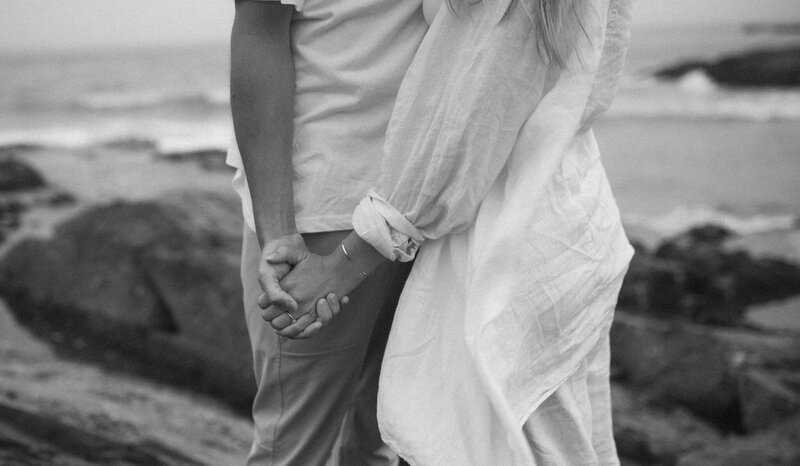 Black and white photograph of a couple standing on the beach, shown from the shoulders down, gently holding hands. One person wears a casual white shirt and pants, while the other wears a flowing long-sleeve dress. Soft ocean waves and rocks are visible in the blurred background.


