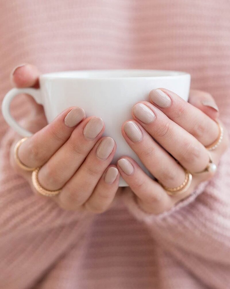 Close-up of a young woman’s hands cradling a warm coffee mug while wearing a pink sweater and gold rings, representing kindness in Xanthe Bookkeeping’s business values.