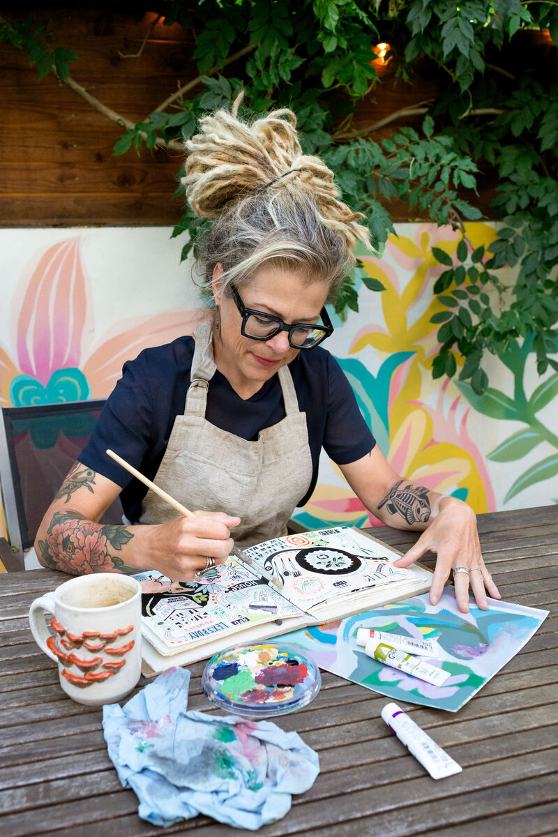 An image of Artist Michelle Allen, taken by Calgary Brand Photography Kate Daniel, looking at her art journal and holding a paint brush.