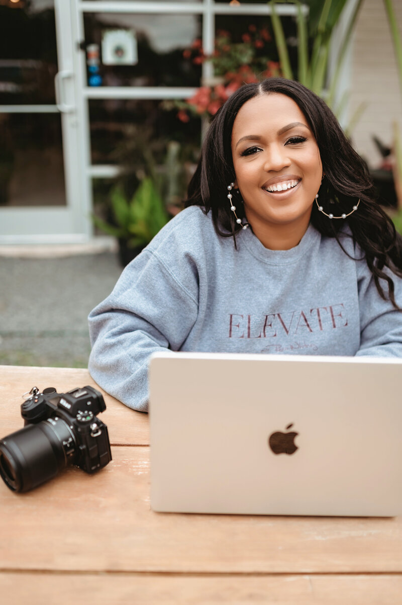 A woman with long dark hair sits at an outdoor table with a laptop and a camera beside her. She is wearing a gray sweatshirt that says "ELEVATE," and she is smiling brightly, with a blurred background of a garden and a building.