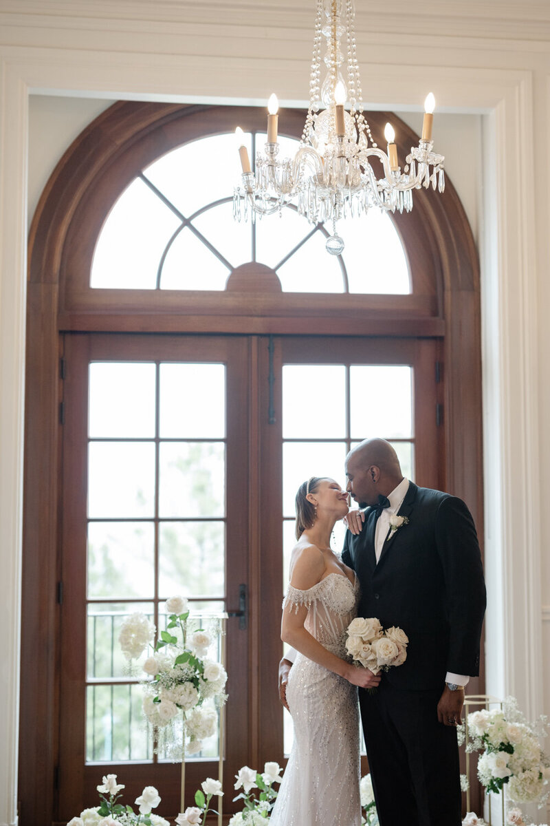 Couple de mariés s’embrassant devant une grande porte vitrée entourés de superbes fleurs blanches, scène romantique et florale d’un mariage raffiné et lumineux