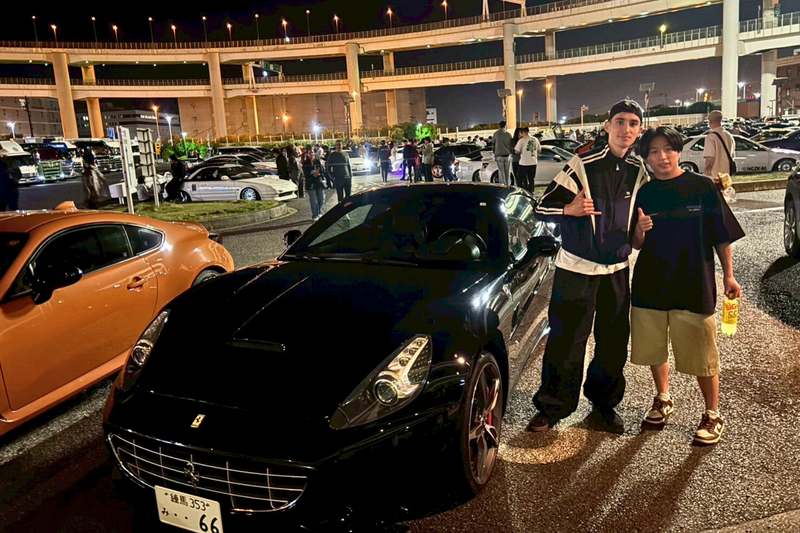 Two guests posing beside a black Ferrari at the Daikoku Parking Area night meet in Tokyo, with car enthusiasts and modified vehicles gathered under elevated expressways.