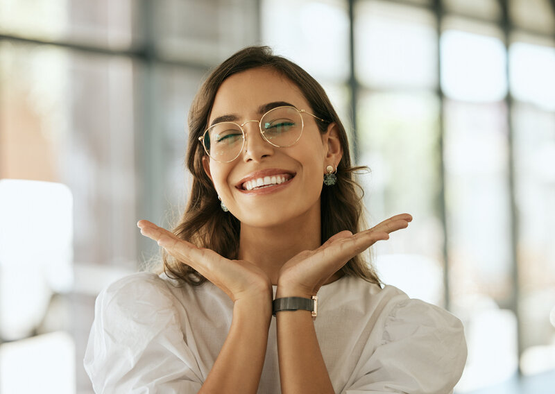 A Latina woman wears glasses and has dark hair. She has a big smile and opens her hands to display her big smile. 