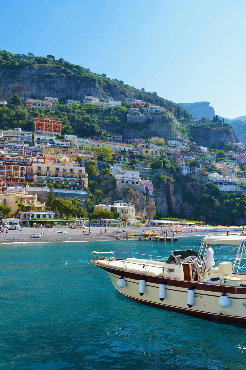 A luxury boat cruises along the turquoise waters of Positano, Italy, with colorful cliffside villas in the background.