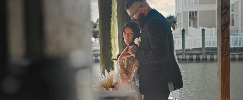 This wedding film frame grab from Films by Brian, a Williamsburg, VA-based destination wedding videographer, shows a beautiful bride holding her groom's hand at a Virginia marina,while looking into the camera.