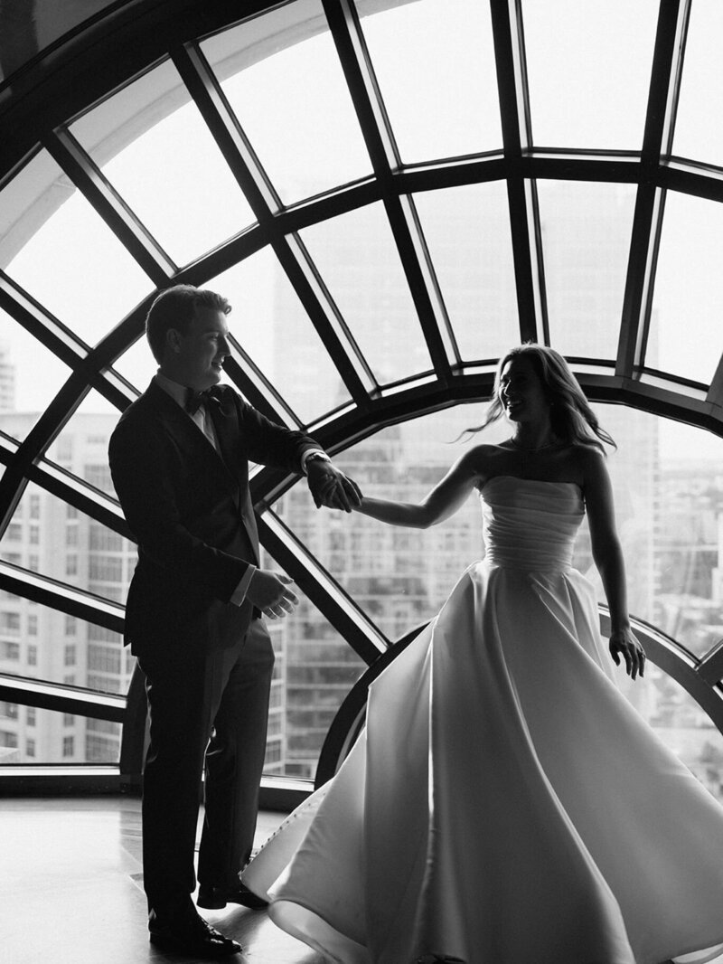 Black and white image of a bride and groom dancing beneath an arched glass window at their elegant city wedding.