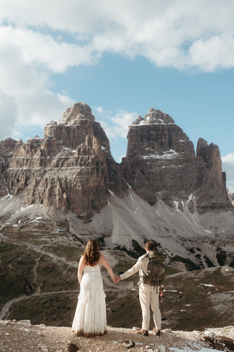 Adventure Elopement Ideas | Eloping couple holds hands standing in front of massive mountain shaped a little like a heart, groom is wearing a hiking backpack