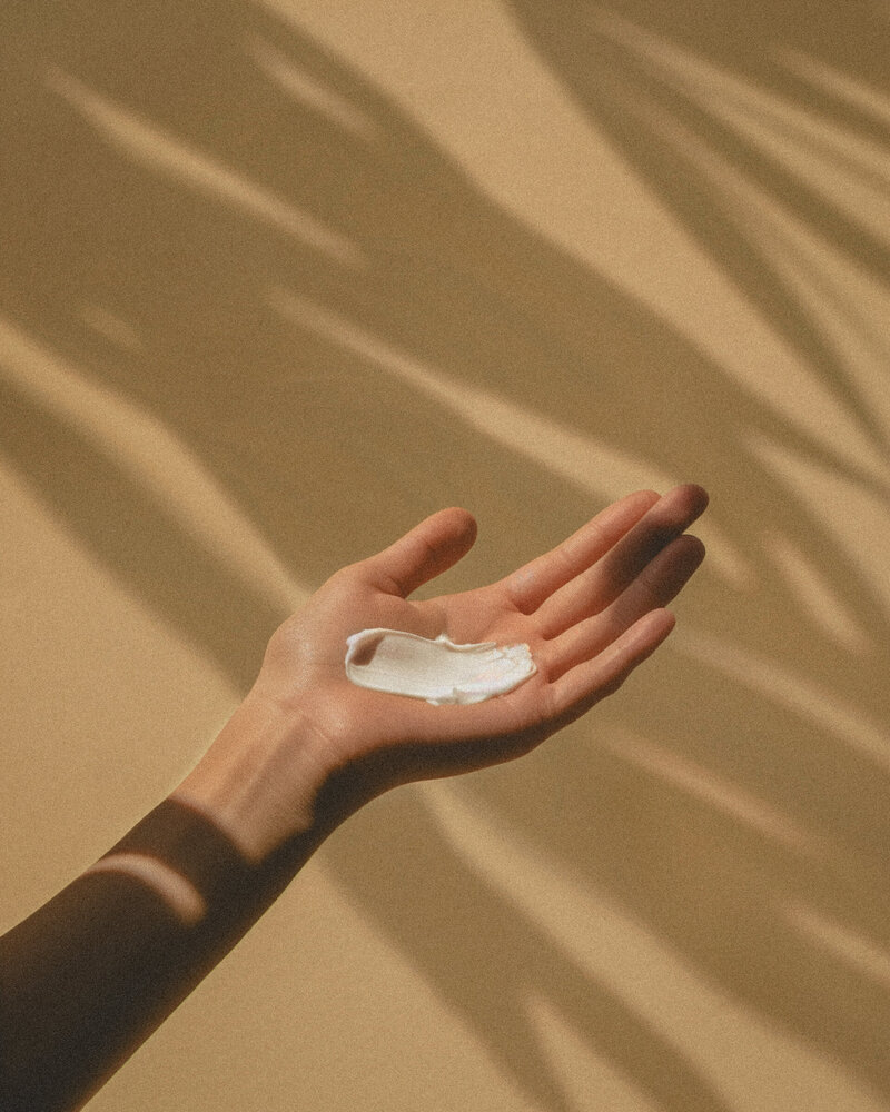 An open hand with skin care cream on the palm is shown against a beige background with soft, shadowed foliage patterns.