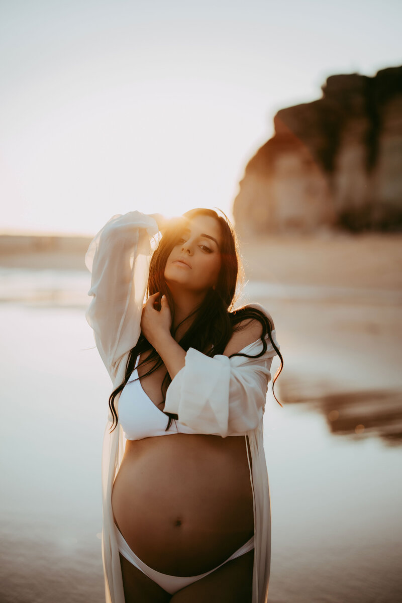 Pregnant woman holding belly at Sydney beach at sunset, photographed by Sophie Lea.