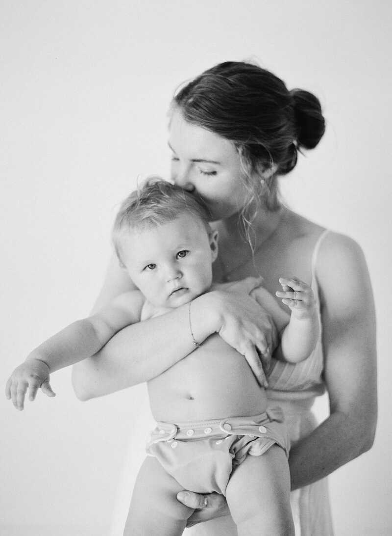 Black and white photo of a mom kissing her six month old son on the head while he looks at the camera