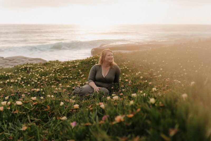Molly, a San Diego photographer, sitting in a field of flowers by the ocean at sunset, enjoying a peaceful moment
