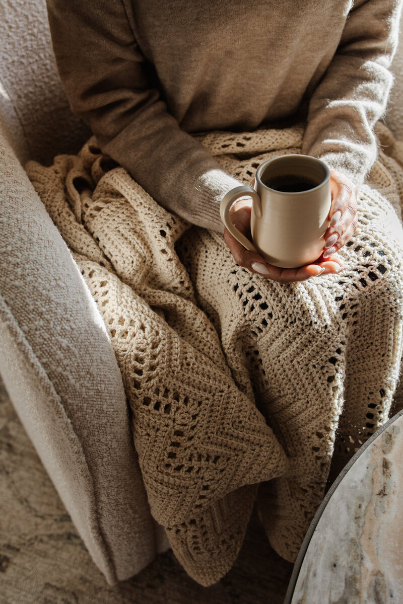 A women sitting in a chair with a blanket on her lap holding a mug of coffee 