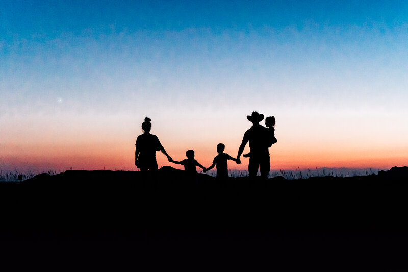 A silhouetted image of Kate Simpson’s family holding hands and walking together against a colorful sunset backdrop, with the father wearing a cowboy hat and holding a young child.