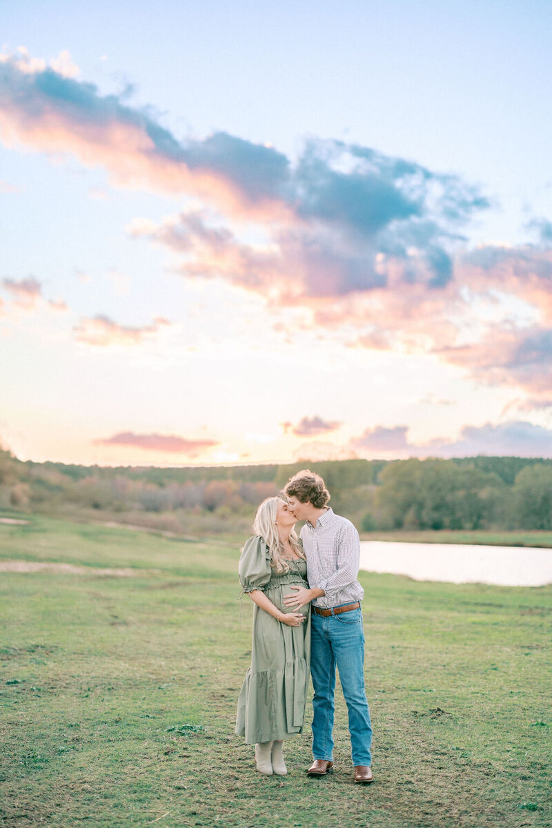 Pregnant woman in green dress kisses husband in field with a pink and orange sunset