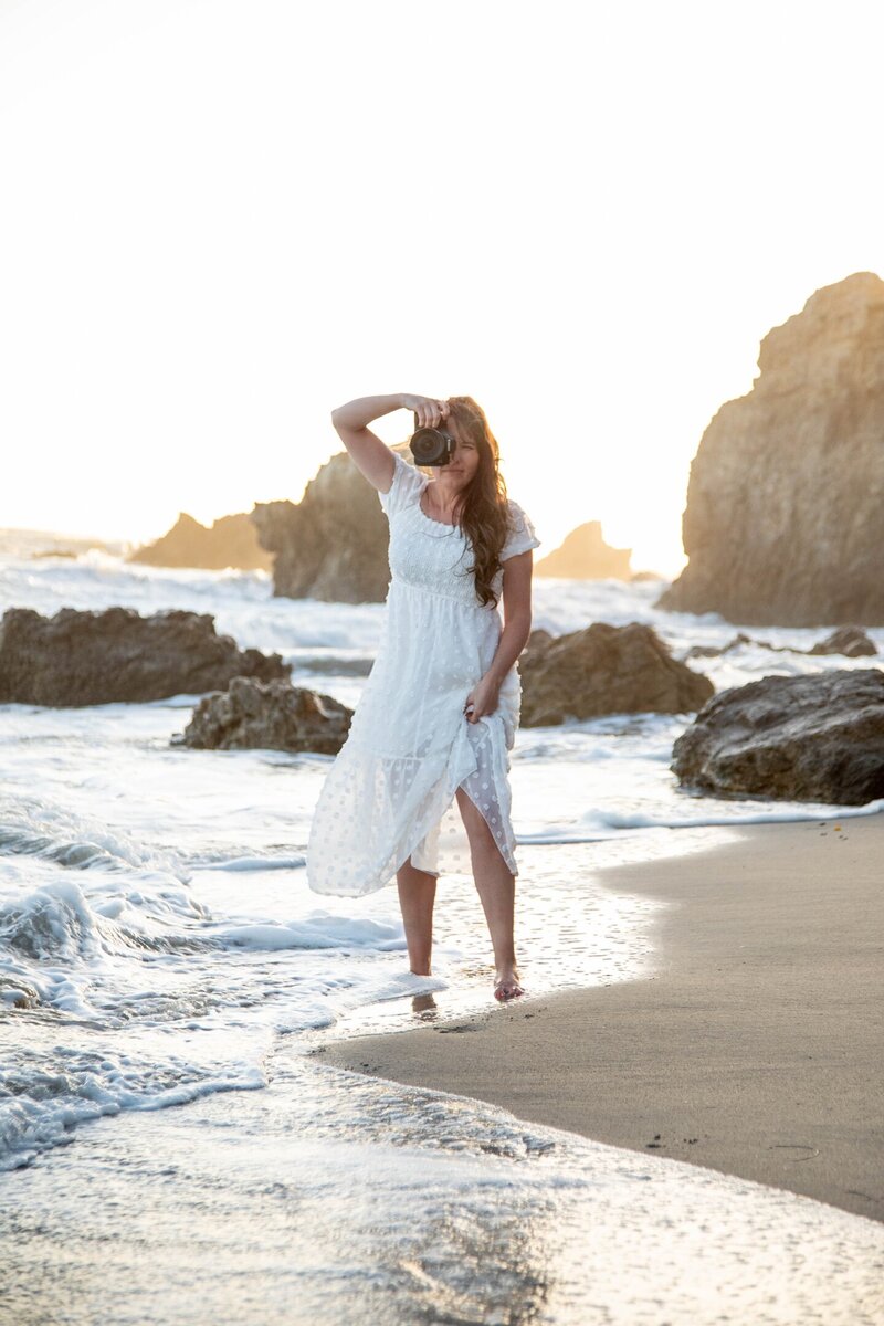 Heidi holding camera up to her face with one hand standing on California beach | Maine Wedding and Portrait Photographer