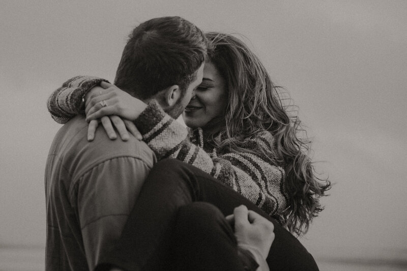 Black and white photo of a sunset session of a couple on Cape Kiwanda Beach. He's holding her in his arms and they're wrapped up close in a moment of windblown passion.