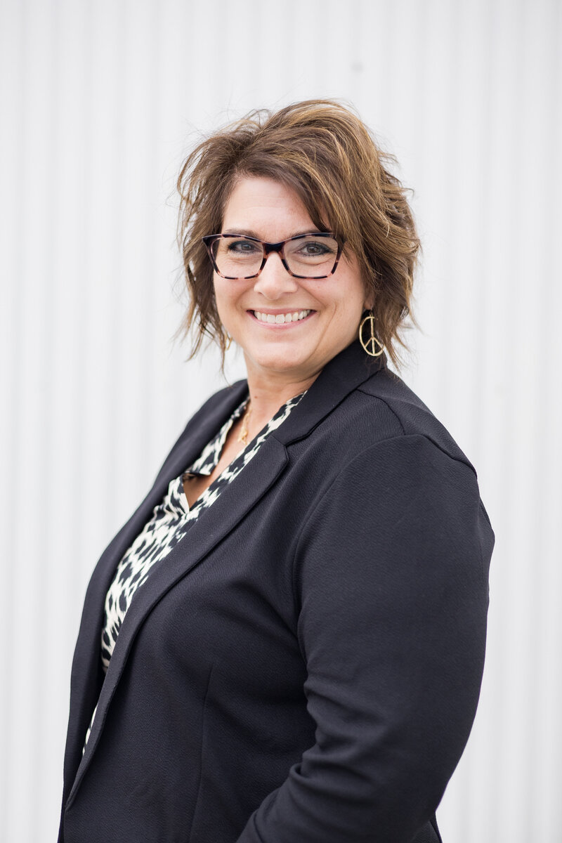 Woman standing in front of a white building with brunette blonde hair, wearing a black dress jacket with a white and black cheetah print shirt, wearing glasses, turned to the side, smiling at the camera