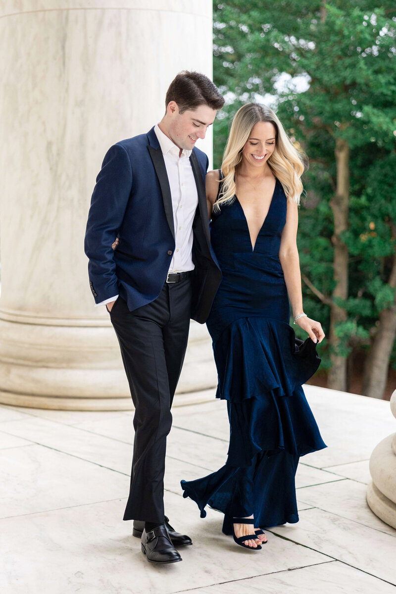 A man and woman dressed formally in blue walk side by side, smiling. The woman wears a sleeveless, deep v-neck gown with ruffles, and the man wears a dark suit with a white shirt. They are outdoors in front of a tall marble column and green trees.