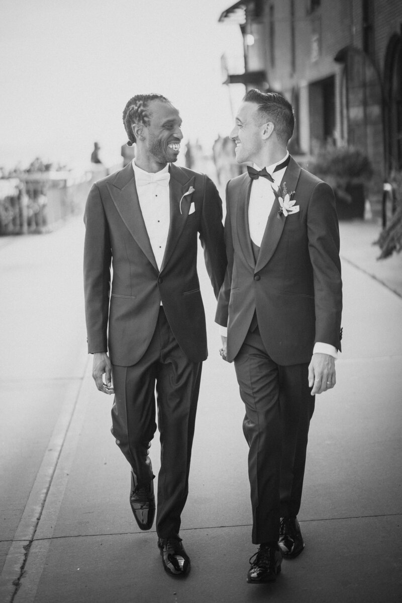 Two grooms holding hands and walking along a sidewalk together.