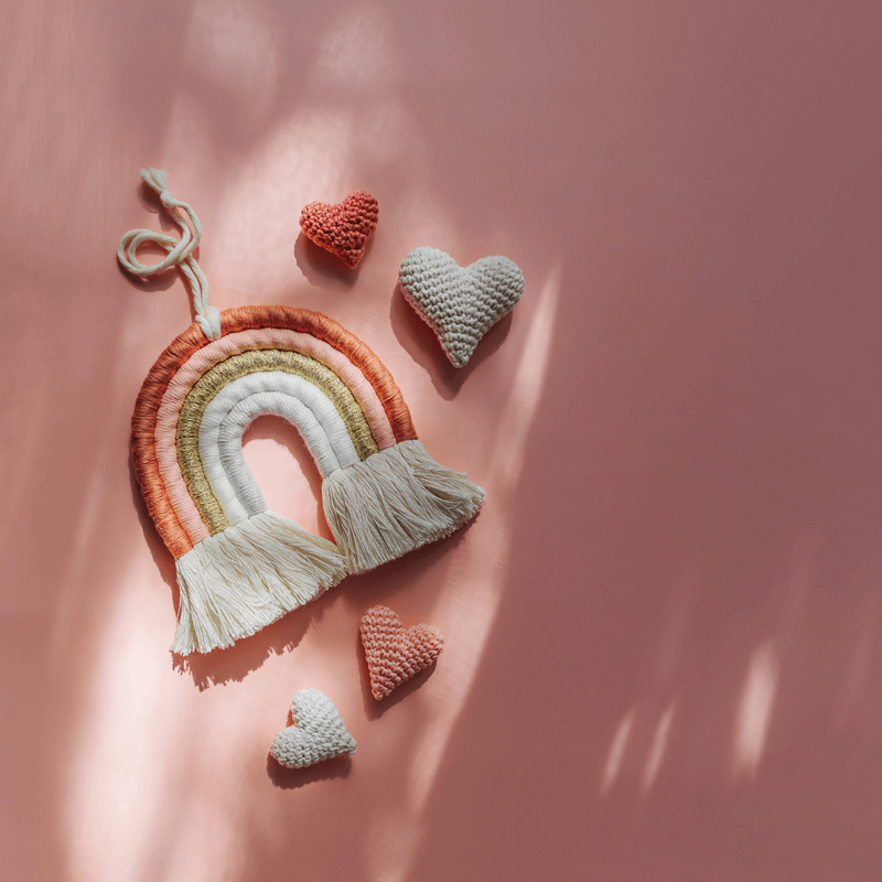 A handmade rainbow wall hanging with a heart on a peach background.