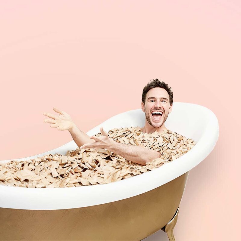 Smiling man in bath filled with soap pods against peach background.