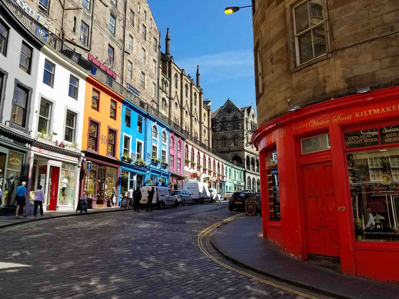 Colorful storefronts line the curving cobblestone street of Victoria Street in Edinburgh, Scotland, United Kingdom, with historic stone buildings towering above under a bright blue sky ©Stephanie Dosch | theViatrix Luxury Travel