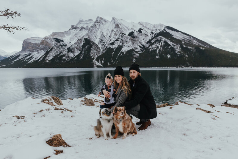 A mother holding her daughter and father crouch beside a mountain lake in the snow  with their two dogs in front of them