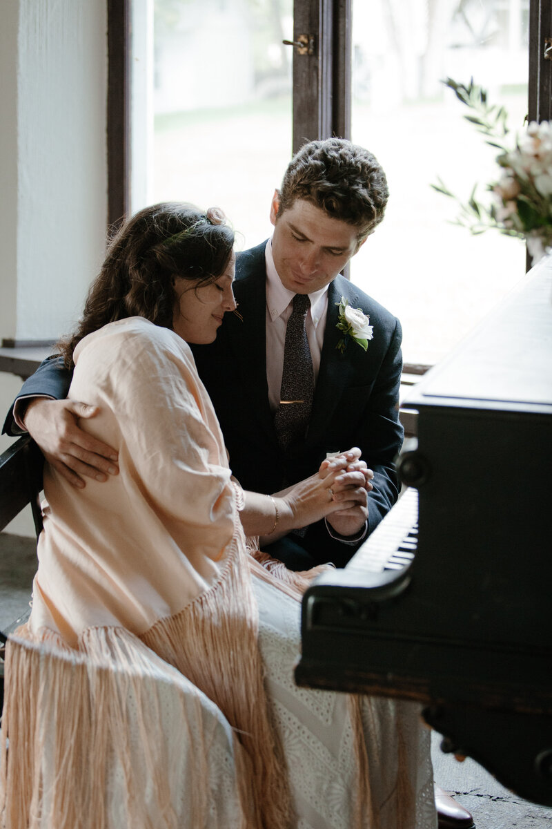 Vintage piano shawl draped over piano as couple embraces on wedding day in VT. Wedding planned by Randi Nonni Events.
