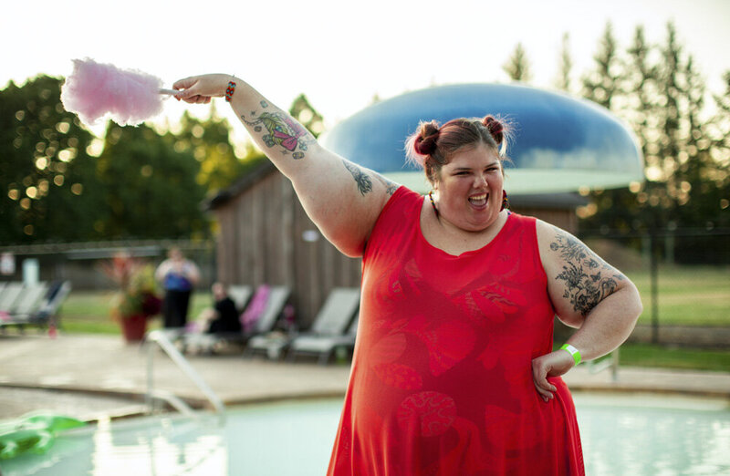 A white, plus-size feminine-presenting person stands outside in front of a pool. They are wearing a red dress over a bathing suit and are holding up a stick of cotton candy, smiling excitedly at the camera. Their hair is in pink space buns, and their arms are covered in colorful tattoos.