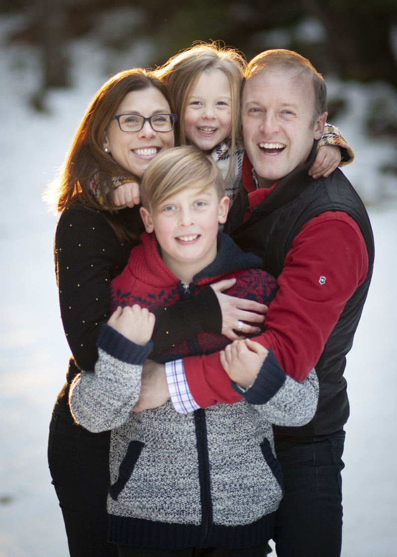 Family of 4 embrace & smile brightly in the snow during a family portrait session in Falmouth, Maine