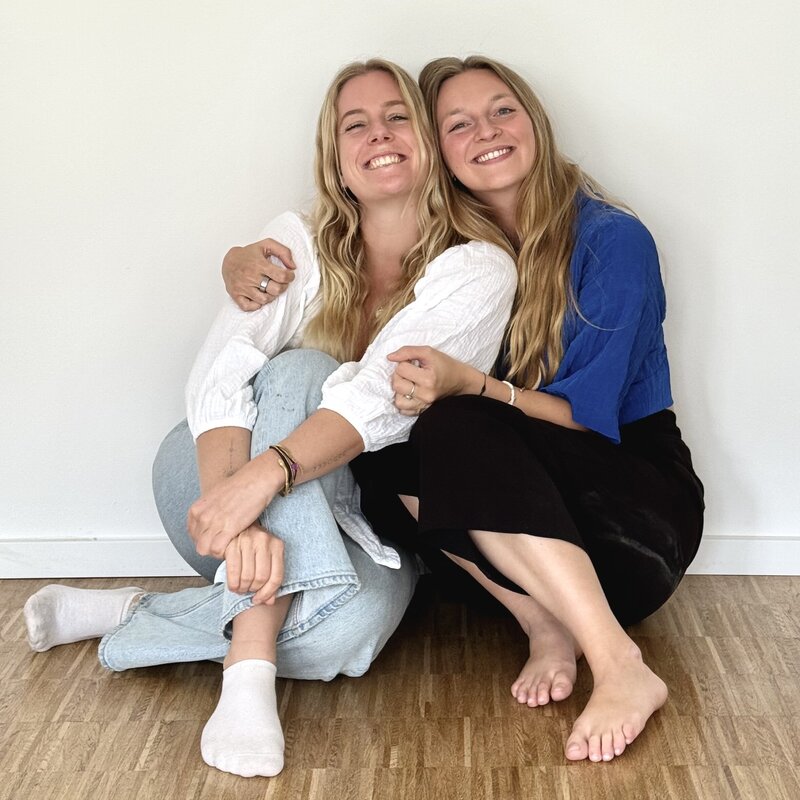 Two women sitting on the floor in front of a wall hugging each other and smiling