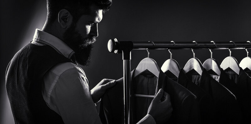 A bearded man in a white shirt and vest studies a suit jacket on a clothing rack in a dark studio, framed by moody lighting.