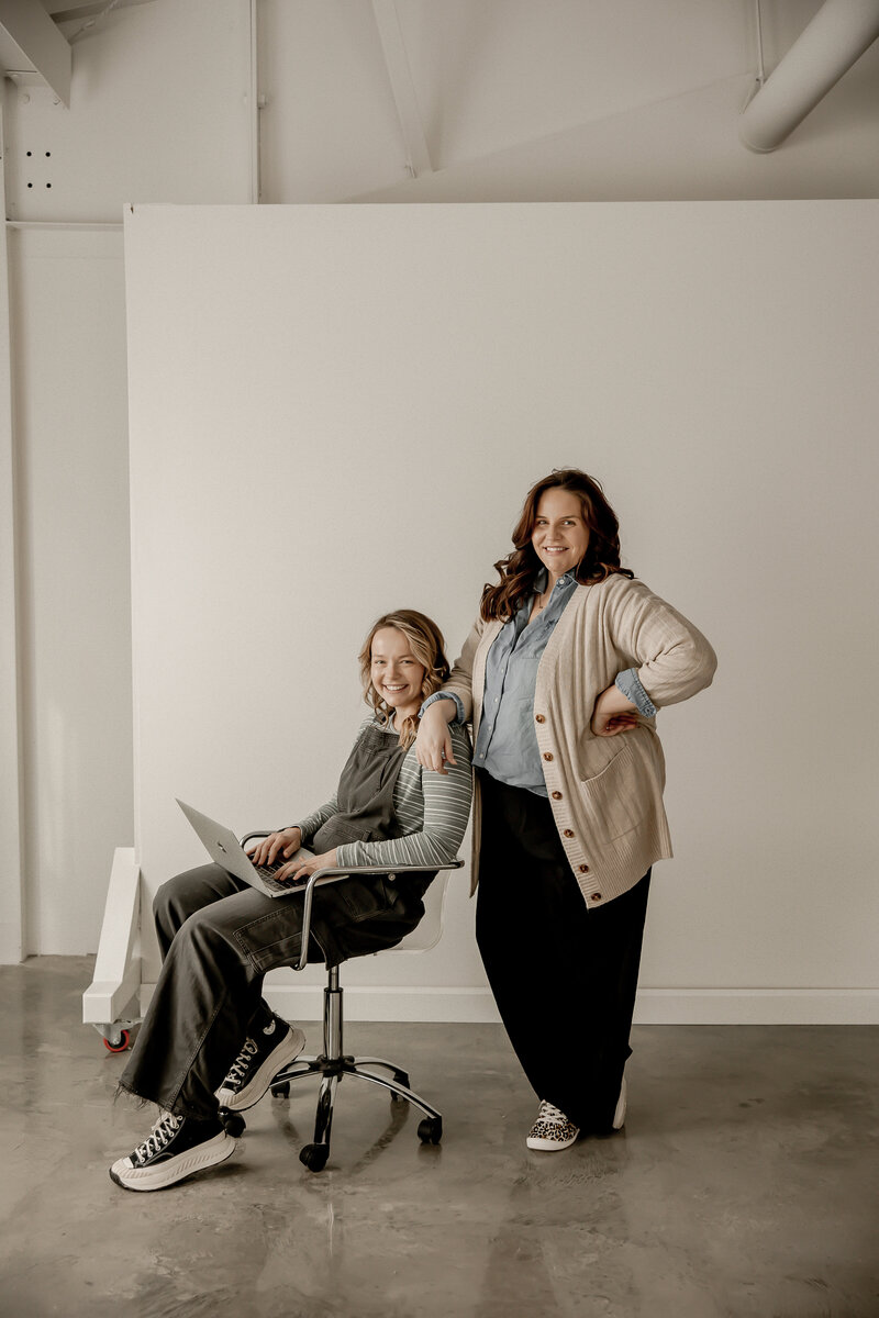 Amber sitting in a chair with computer on lap while lacey stands leaning on. her shoulder