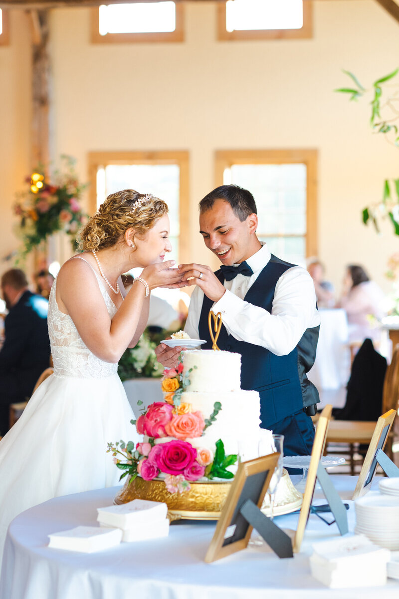 A bride and groom standing in front of their wedding cake on their wedding day. The wife has just taken a bite of the cake, fed to by the groom, and is smiling.