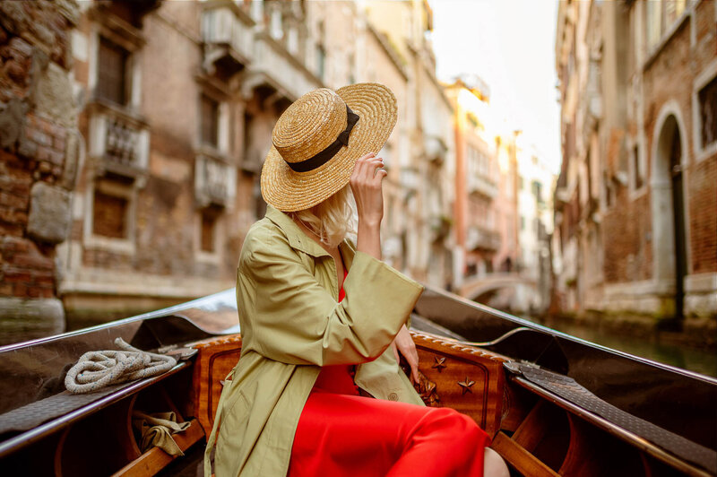 Person wearing a straw hat and sitting in a gondola on a canal in Venice, surrounded by historic buildings.