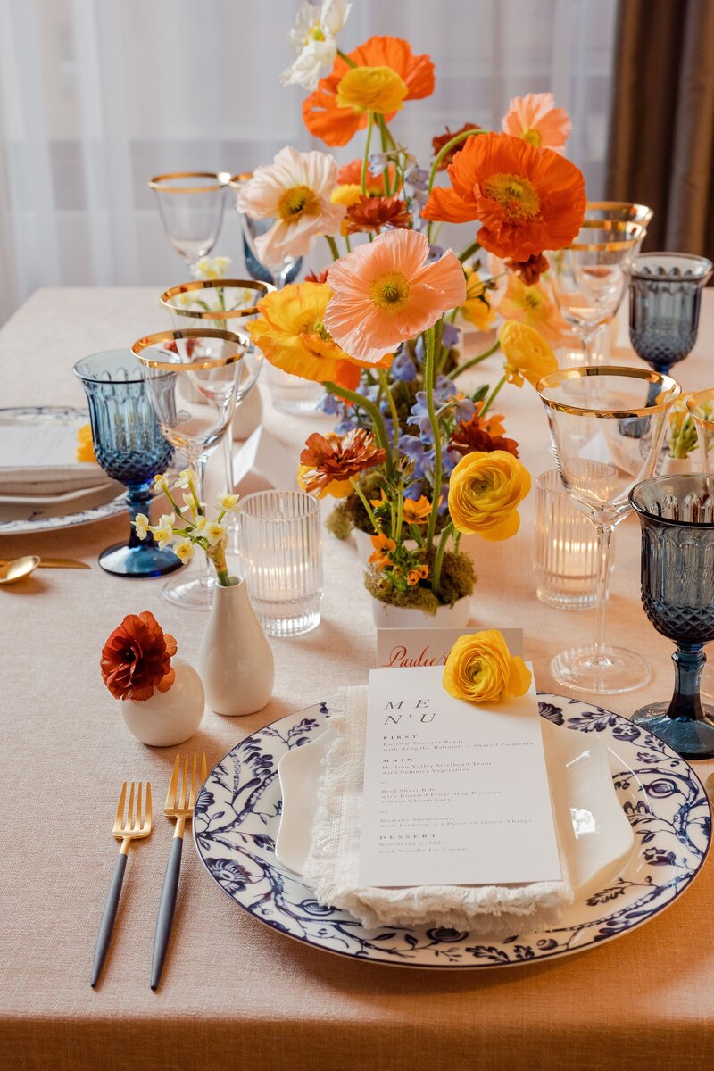 Elegant wedding table setting featuring vibrant orange and yellow flowers, blue goblets, gold flatware, and a beautifully designed menu.