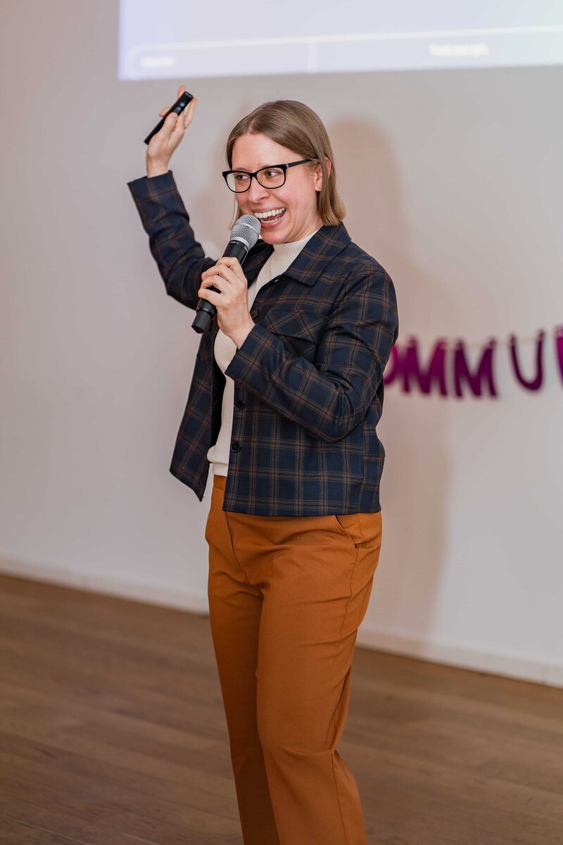 Stephanie wears a navy-blue jacket and burnt orange pants while speaking in front of a group.