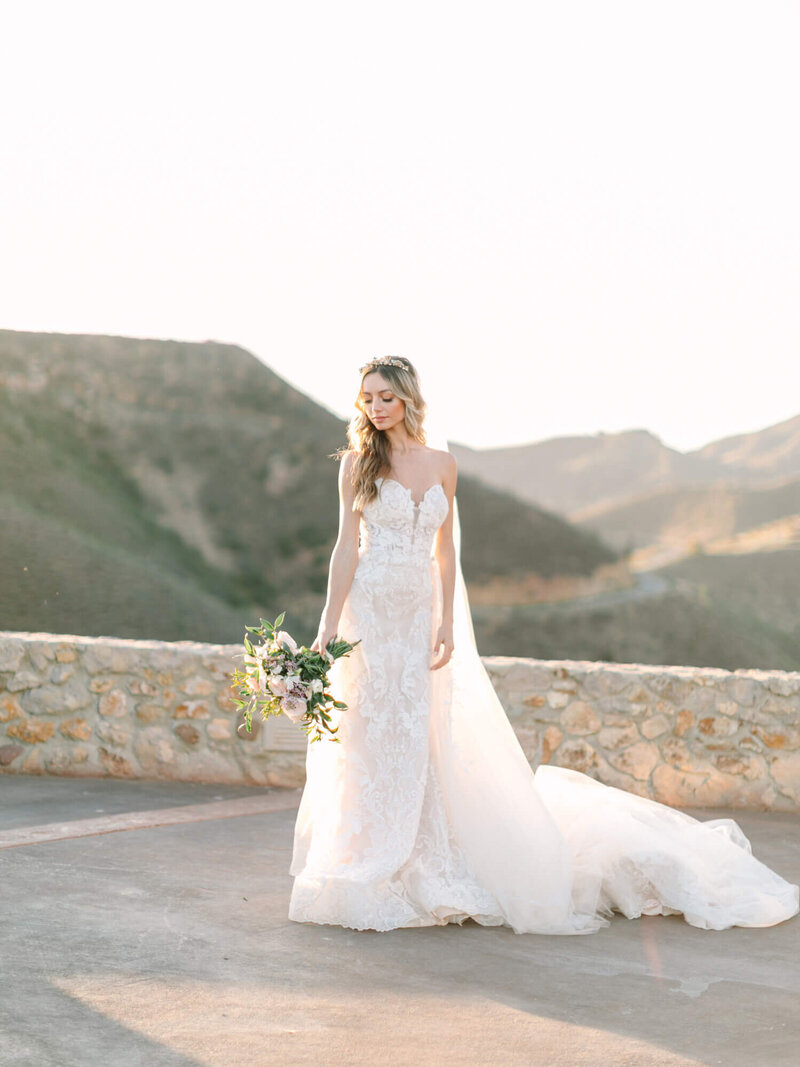 beautiful dreamy bride in a lace gown and flowing train stands outside on a sunny day, holding a bouquet with mountains in the background.