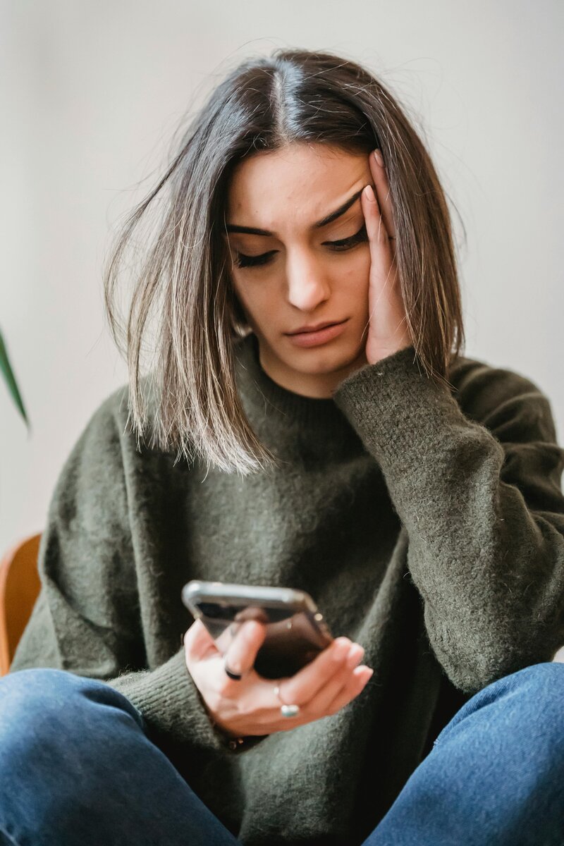 A young woman with shoulder-length dark hair sits cross-legged in a sweater and jeans, holding a phone in one hand and resting her head in the other.