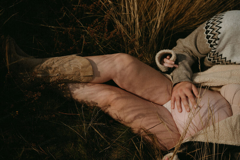 Femme allongée dans l'herbe avec la main de son compagnon posée sur son ventre rebondi. Shooting photo grossesse vers Rennes en Bretagne par Kamescop.