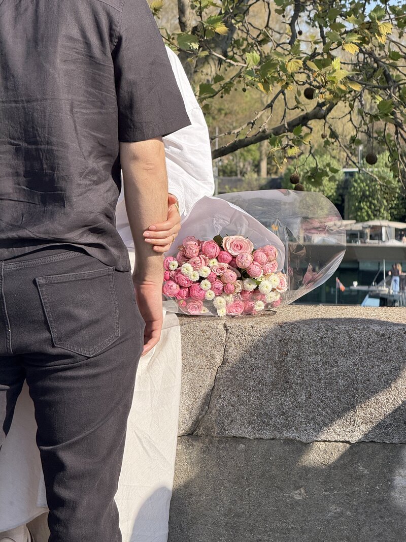 A person in black holds another's hand on a sunny day by a stone wall. A bouquet of pink and white roses rests nearby, under a tree with leaves.