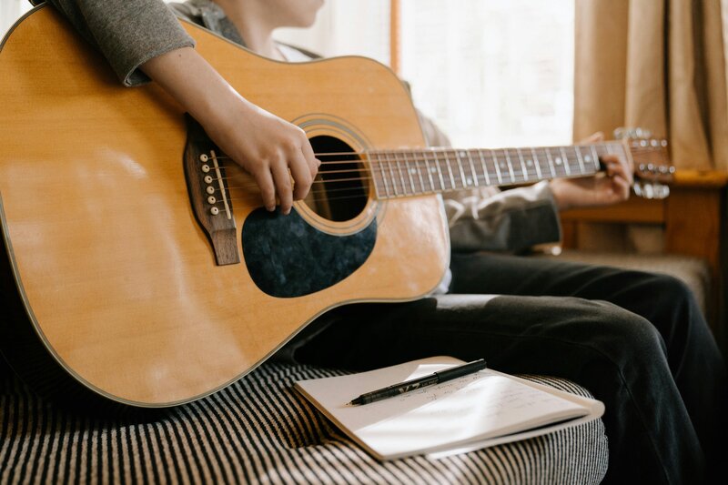 Individual sitting on a couch playing an acoustic guitar with a notebook and pen. Songwriting