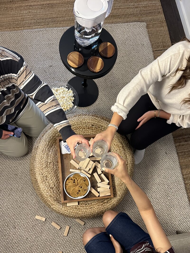 Three people toast with glasses over a woven ottoman holding cookies and Jenga blocks. Popcorn and a water pitcher are nearby, creating a cozy, playful vibe.