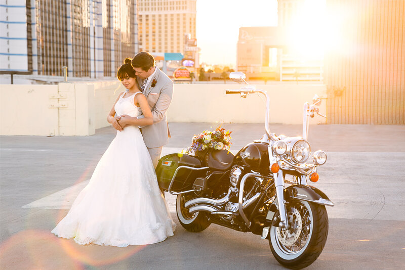 Bride and Groom embrace on a Las Vegas Rooftop next to their Harley Davidson motorcycle at sunset with the sun creating a beautiful backlight on the couple and the bouquet sitting on the motorcycle.