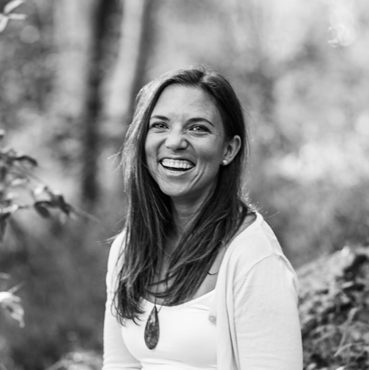 Women in golden light, wearing dark rimmed glasses and striped shirt, smiling down at her hands as she rolls out clay