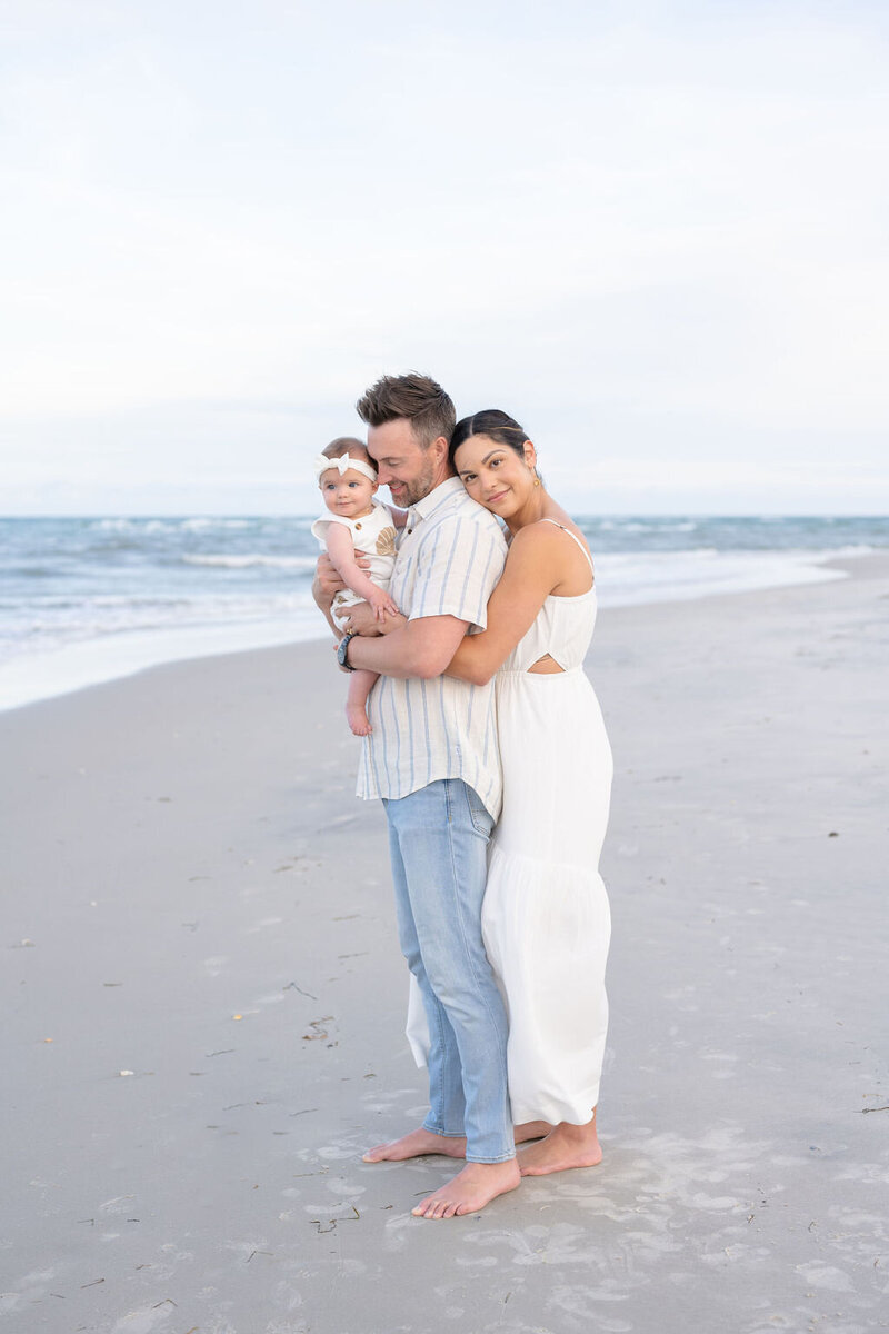 father holding a baby girl wearing a white bow on north topsail beach as mother hugs his back and rests her head with the tide is coming in and the sky is white and clear