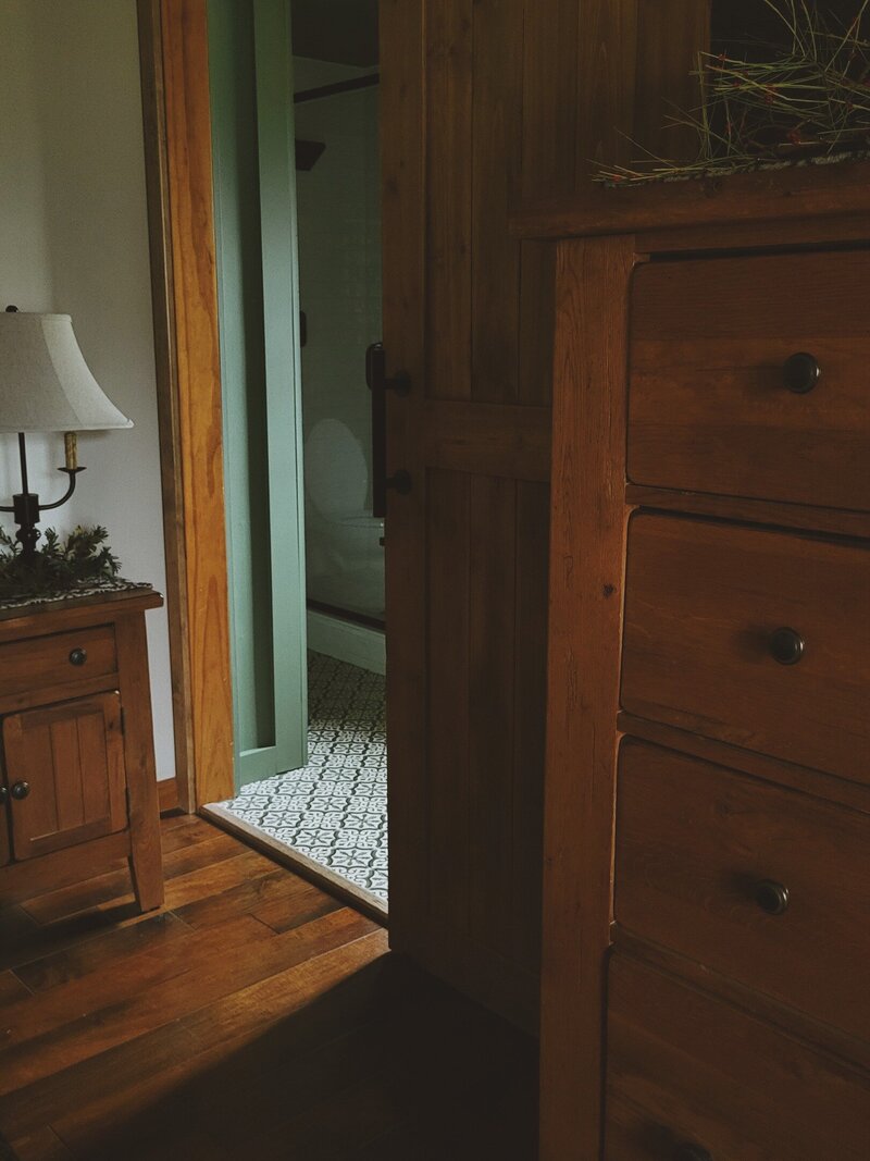 Wooden bedroom floor with oak bureau, and nightstand wtih candelabra lamp, with a peek into the bathroom where you can see the patterned tile floor through the matching wood stain sliding door