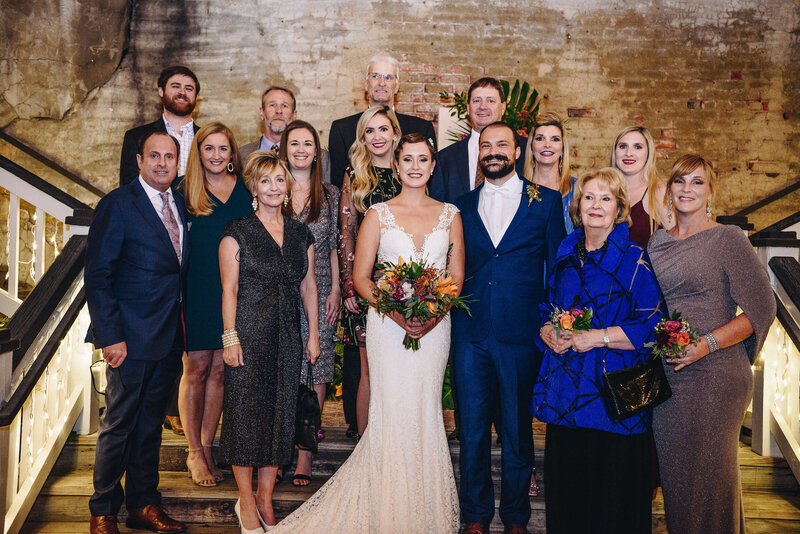 A wedding group photo featuring the bride in a white gown and the groom in a blue suit at the center, surrounded by smiling family members on a staircase with exposed brick walls in the background.